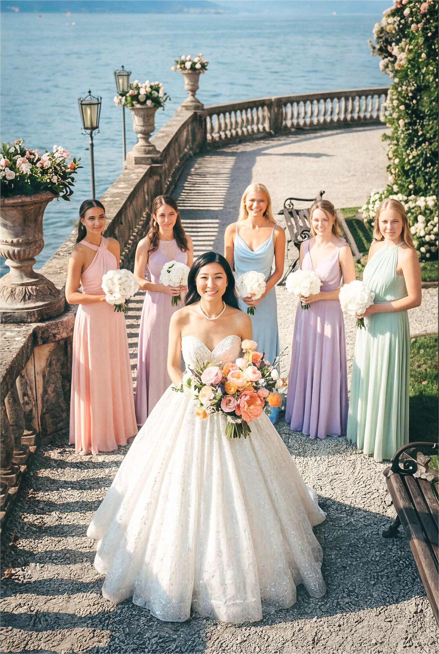 Bridal party in elegant pastel dresses by a scenic lake, surrounded by blooming flowers and classic architecture.