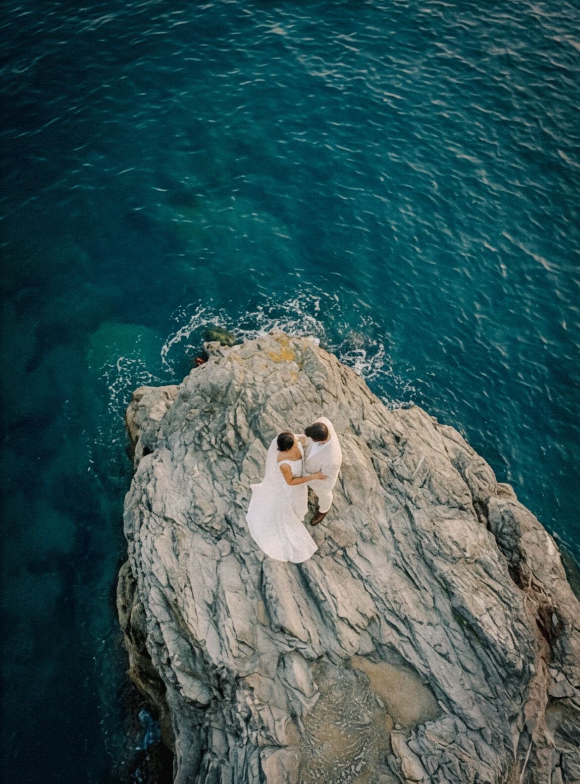 Couple embracing on a rocky outcrop by the serene blue ocean during their romantic destination wedding.