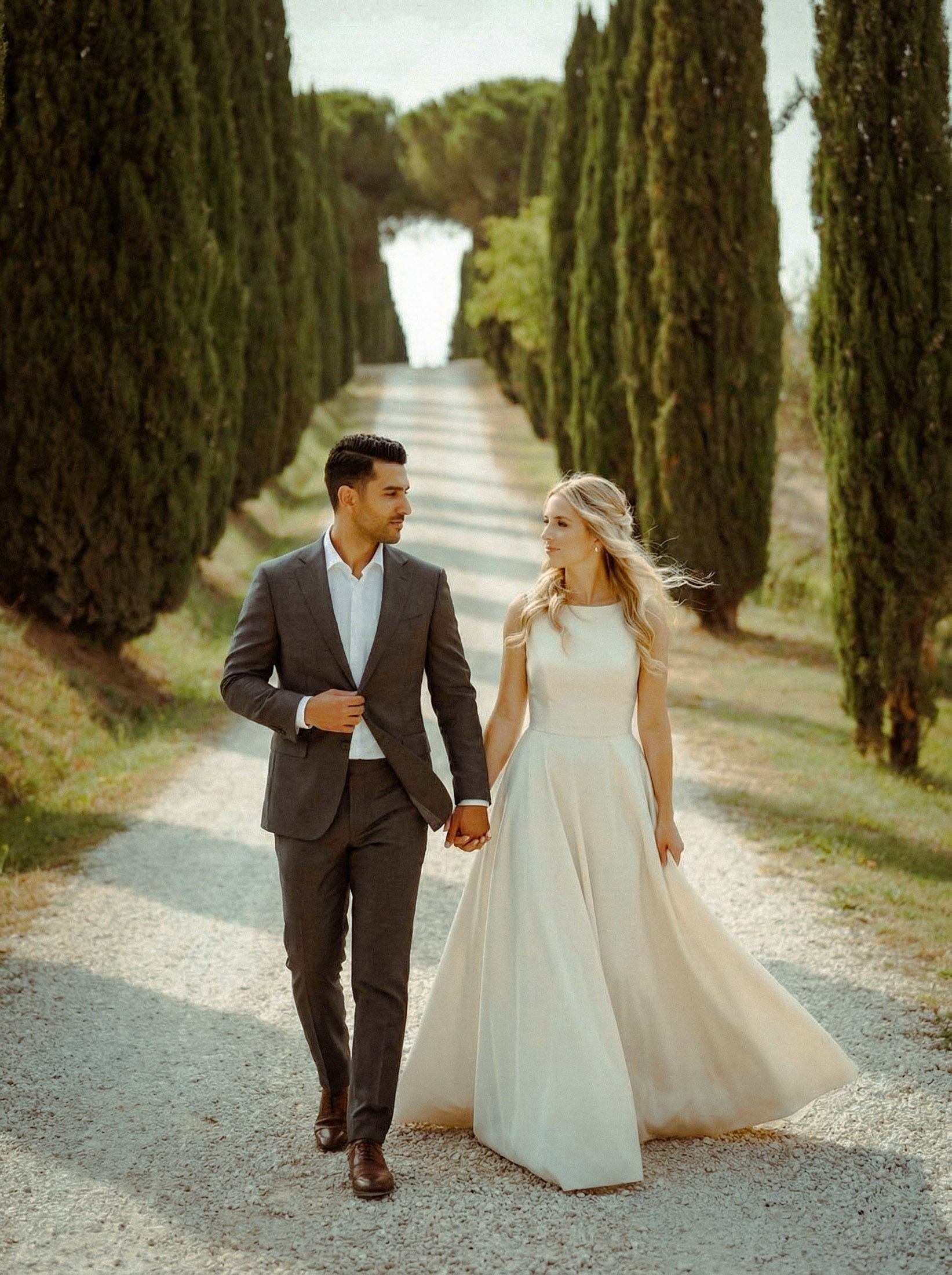 Couple in elegant attire walking hand-in-hand down a scenic path lined with tall cypress trees, exuding romance.