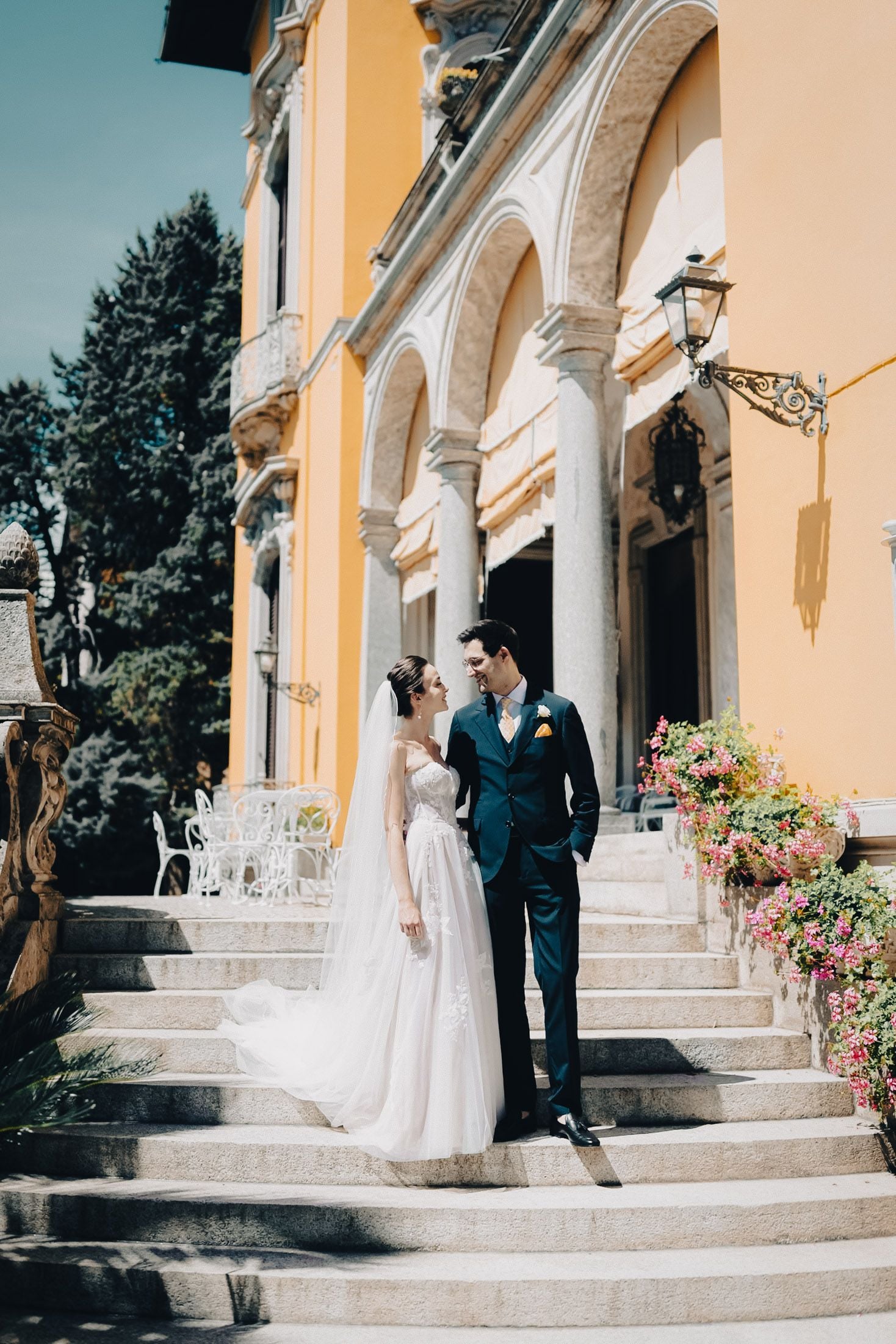 A couple in wedding attire standing on stone steps of a vibrant villa, surrounded by greenery and flowers, exuding romance.