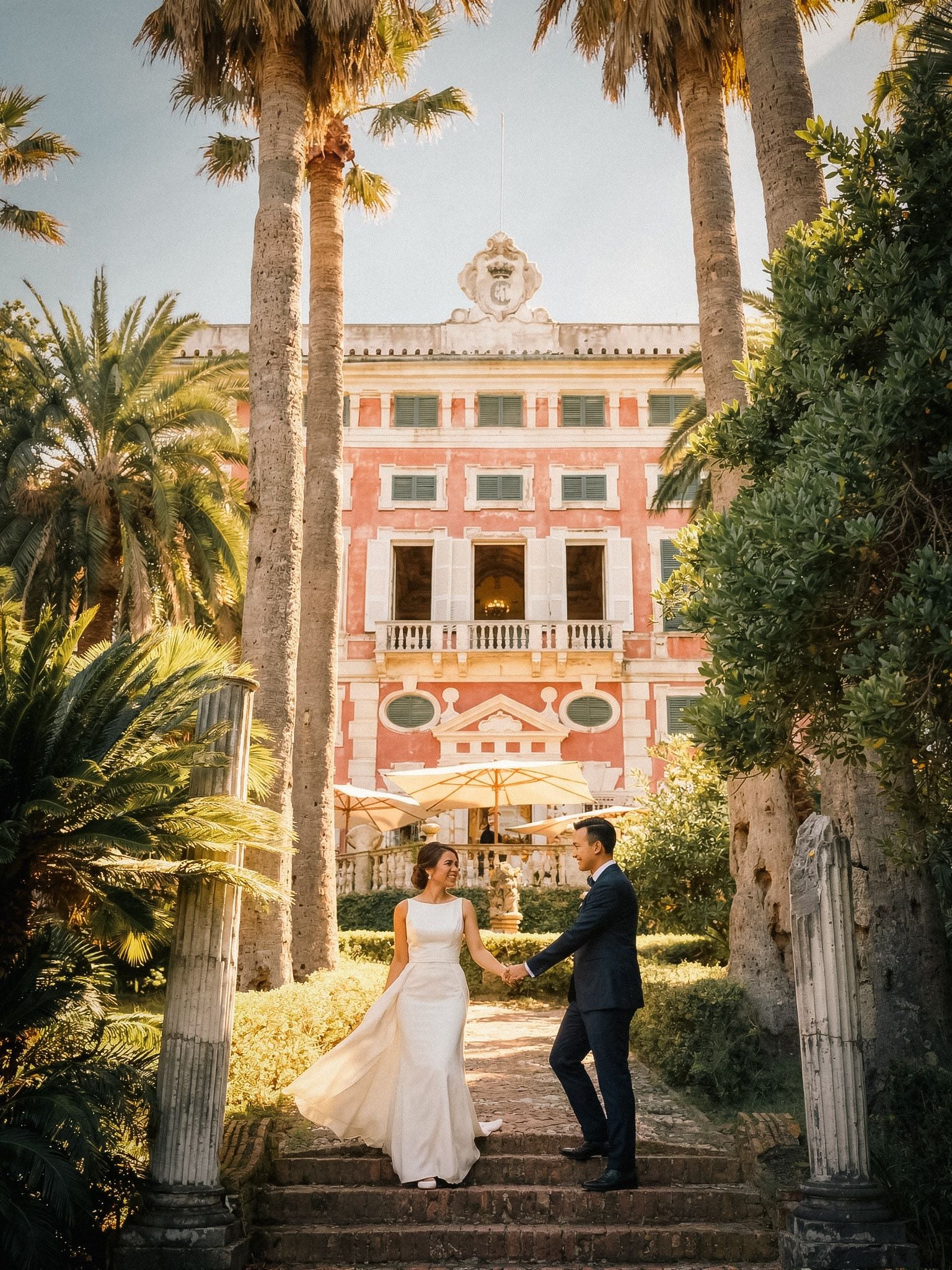 A joyful couple holding hands on stairs, surrounded by lush palms and vibrant pink villa, evoking a romantic atmosphere.